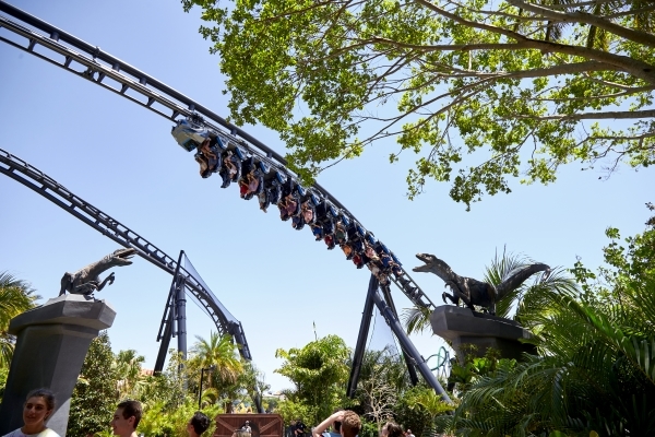 Riders a black roller coaster track hang upside down surrounded by two velociraptors on pillars.