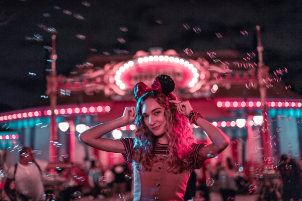 A woman wearing Minnie Mouse ears stands in front of the red and pink lit Dumbo attraction at night with bubbles floating in the background. 