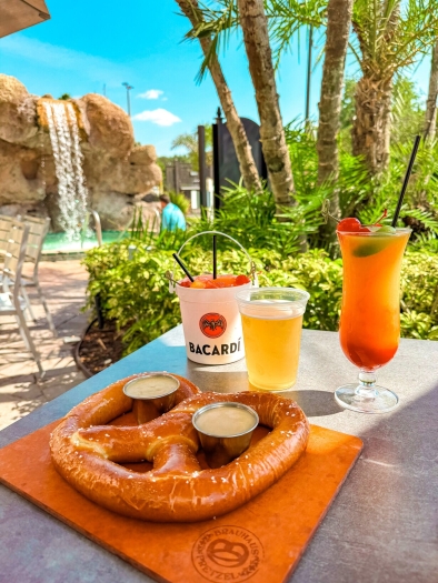 A large soft pretzel on a wooden platter, a beer, an orange cocktail, and a drink in a bucket that says "Bacardi" sit in front of a garden with palm trees, and a water fountain in the background. Rosen Plaza is close to all of the best food halls in Orlando. 