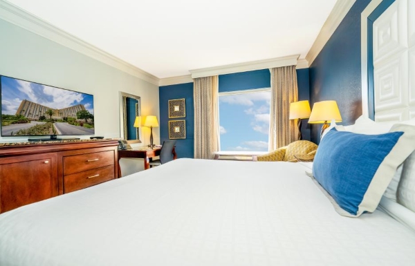 A hotel room with a dark blue accent wall, a bed with white linens and a blue pillow, and a wood dresser with a television. Rosen Plaza is a wonderful destination for solo travel.