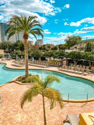 The deck surrounding a crystal blue pool with palm trees and blue sky. A pool day at Rosen Plaza is one of our favorite things to do on Thanksgiving in Orlando.