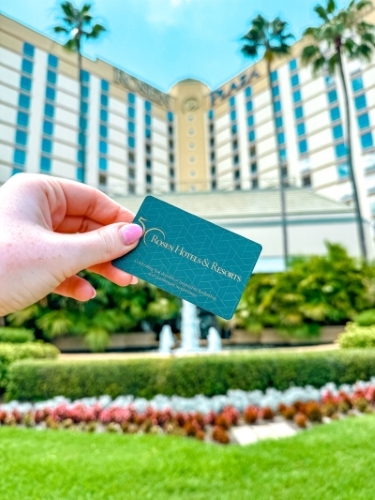 A hand holding a Rosen Hotels & Resorts room key in front of Rosen Plaza hotel. We're the perfect place to stay if you're attending any events at the OCCC this year.