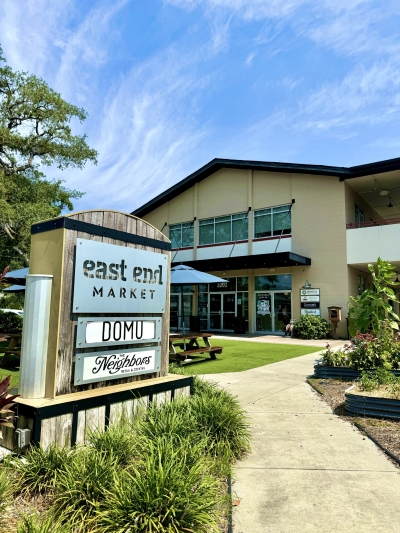 A sign reads "East End Market" in front of a two-story building surrounded by lawn, gardens, and pathways. East End Market is an incredible food hall in Orlando. 