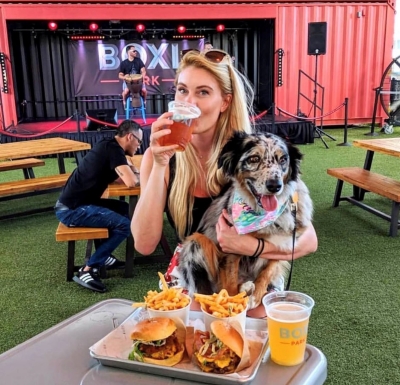 A blonde woman sips a beer with a gray and black dog in her lap. Behind her is a stage with a sign that reads "Boxi Park" and in front of her is a tray of food with burgers, fries, and beer. Boxi Park is a great Food Hall in Orlando. 