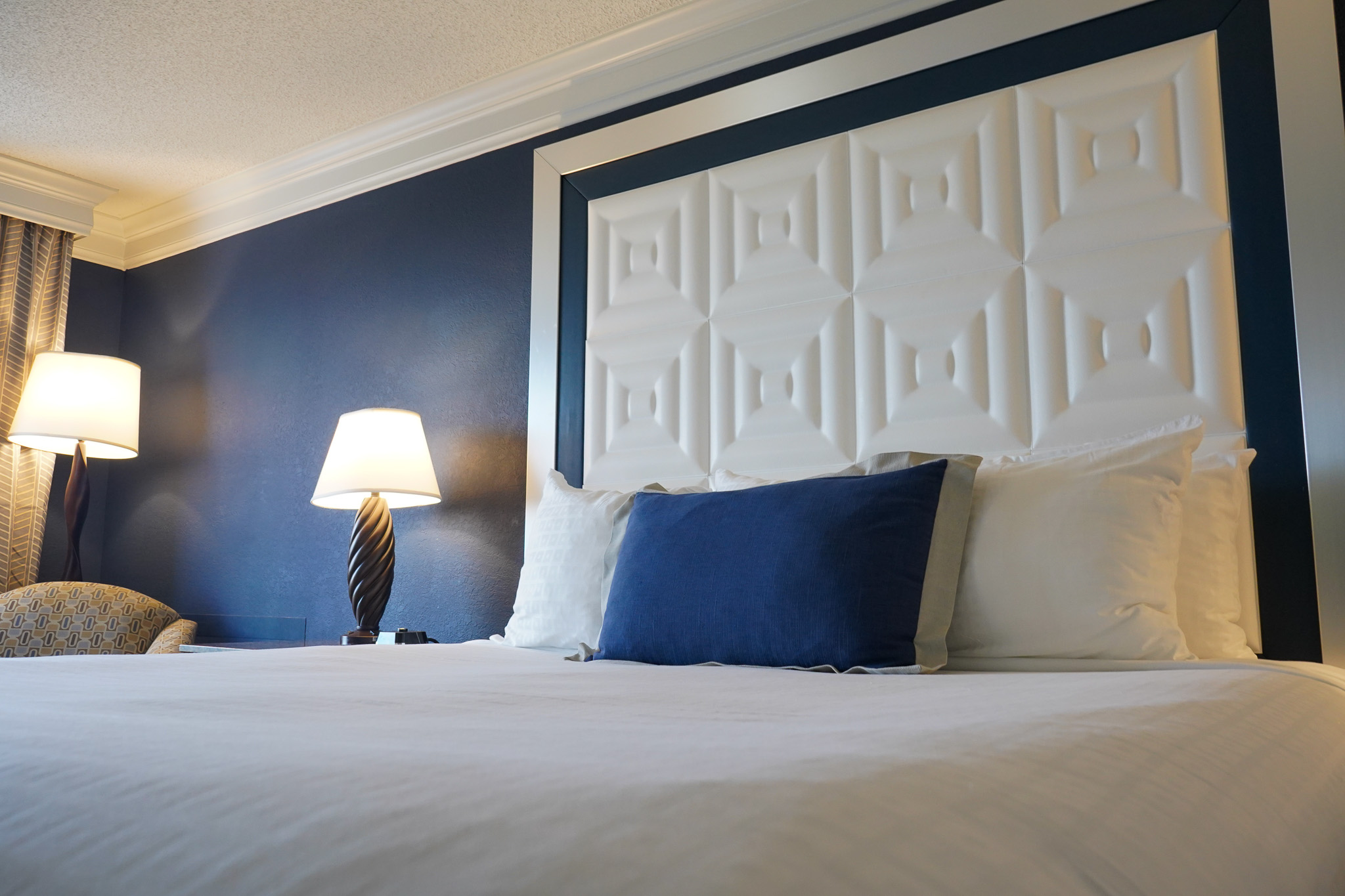 Close-up of a made-up hotel bed with white sheets, pillows, and a blue accent pillow against a dark blue wall at Rosen Plaza in Orlando.