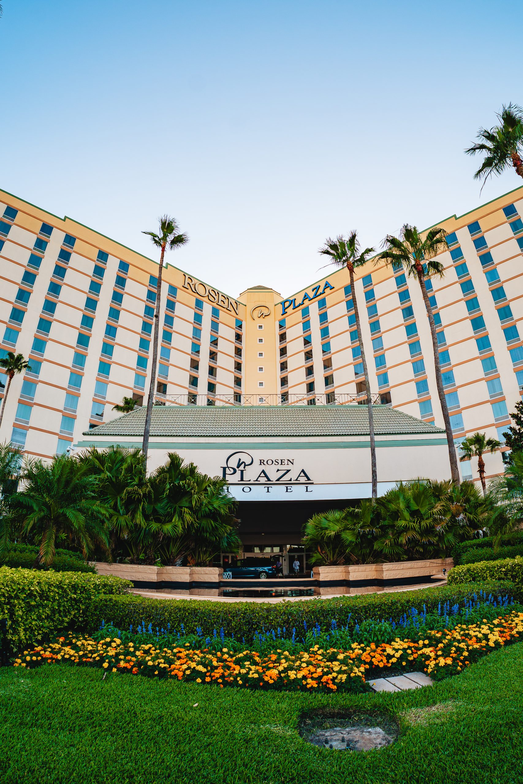 A view of the Rosen Plaza Hotel with its curved facade, prominent signage, and tropical landscaping including palm trees and flowering bushes.