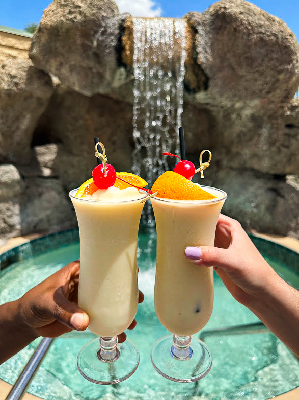 Two frosty piña coladas garnished with cherries and oranges, held against a backdrop of a rock waterfall cascading into a turquoise pool at Rosen Plaza in Orlando.