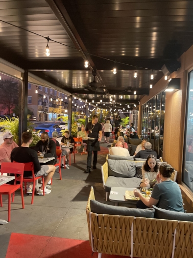 A covered patio at night with booths and tables of people, string lighting overhead, and waiters walking through the aisle. The Marketplace in Avalon Park is a great food hall in Orlando.