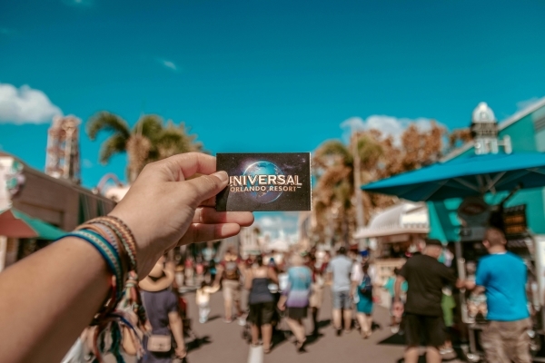 A left hand with bracelets holds a Universal Orlando Resort ticket up against a background of crowds, attractions, and palm trees. 