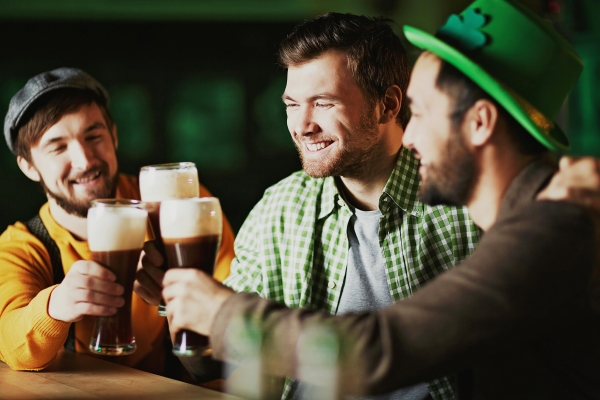 Three smiling men sitting at a bar toast with foamy pints of dark beer.