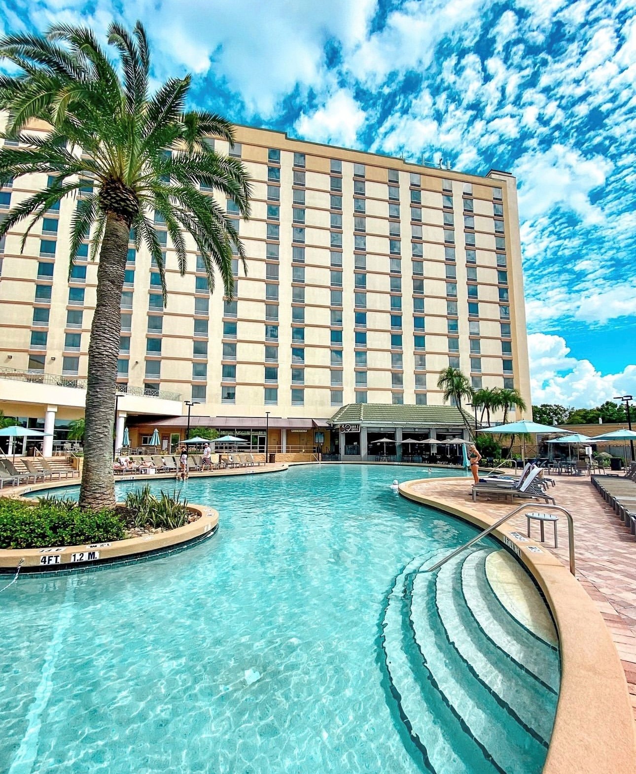 The Rosen Plaza Hotel with a large swimming pool in the foreground, featuring steps leading into the water and palm trees along the edge. Taking an evening dip in the pool is the perfect way to make the most of daylight saving time in Orlando.