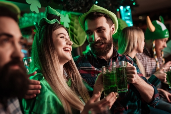 In a small crowd, a smiling man and woman in St. Patrick’s Day green toast two green beers. 